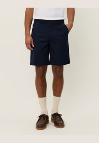 Pantaloni scurți de culoare bleumarin, dintr-un material neted, cu buzunare laterale, închidere cu buton și o croială clasică, asortați cu șosete de un crem deschis și pantofi maro.