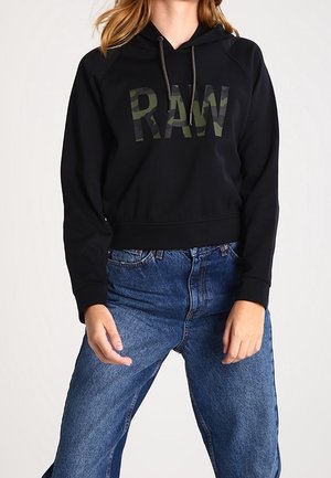 Kobieta w czarnej bluzie z kapturem z napisem "RAW" w kamuflażowym wzorze i niebieskich jeansach z wysokim stanem stoi z rozluźnionymi rękami.