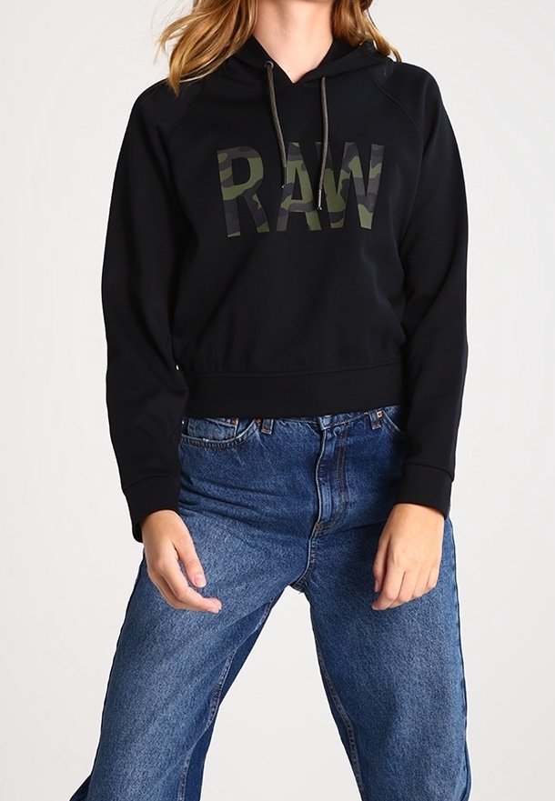 Femme portant un sweat à capuche noir avec "RAW" en imprimé camouflage et un jean bleu taille haute, debout les mains détendues.