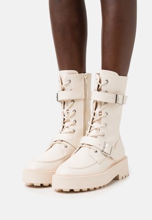 Beige nahkarivi saapaat, joissa on nauhoitus etupuolella, soljikiinnitykset ja tukevat pohjat. Ominaisuuksiin kuuluu sileä pinta ja kestävä rakenne.