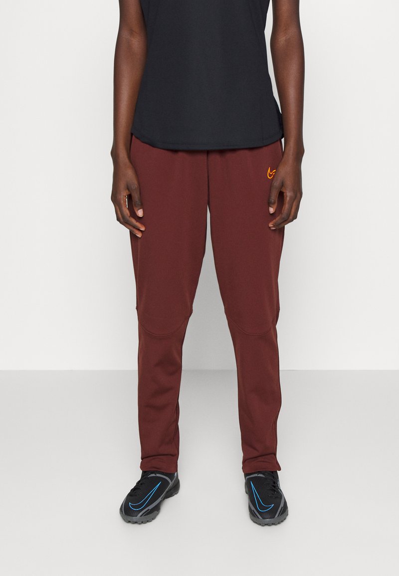 Bordeaux sportbroek met een losse pasvorm, met een oranje logo op de voorkant en vochtafvoerende stof. Zwarte schoenen met blauwe accenten.