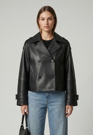 Kvinde iført en sort dobbeltradet læderjakke og blå jeans, der holder en sort håndtaske mod en ensfarvet grå baggrund.