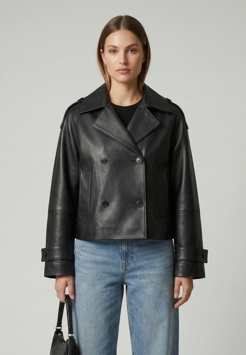 Frau trägt eine schwarze, doppelreihige Lederjacke und blaue Jeans und hält eine schwarze Handtasche vor einem schlichten grauen Hintergrund.