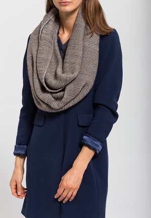 Femme portant un manteau bleu marine et une écharpe tubulaire en tricot taupe épaisse et texturée, les mains détendues le long du corps.