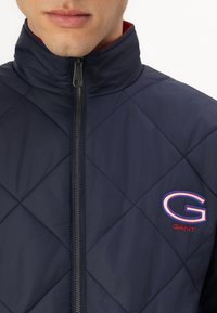 Mann trägt eine marineblaue gesteppte Jacke mit Stehkragen und sichtbarem Frontreißverschluss, die ein großes gesticktes "G" und das "GANT"-Logo auf der Brust zeigt.