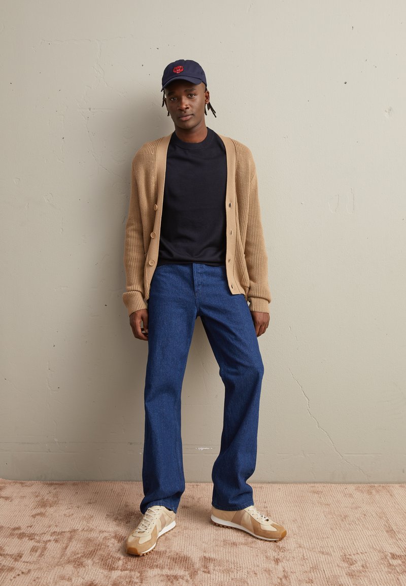 Jonge man die een marineblauwe pet, zwart T-shirt, beige cardigan, blauwe spijkerbroek en beige sneakers draagt, staat tegen een licht gestructureerde muur en een vloerbedekking.