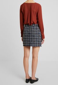 Blouse orange brûlé associée à une jupe en tweed multicolore, dotée d'une fermeture éclair et d'un ourlet à franges, sur un fond clair.