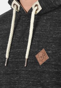 Hoodie gris foncé avec un tissu texturé, un cordon de serrage en cuir crémeux et une étiquette en cuir marron ornée de décorations.