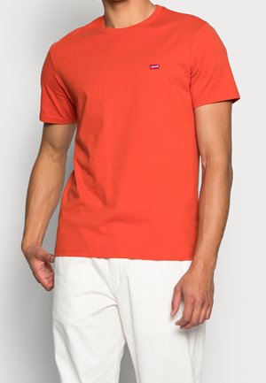 Mężczyzna ubrany w prostą czerwoną koszulkę Levi's z krótkim rękawem i małym logo na piersi, w białych spodniach, stoi na jednolitym tle.