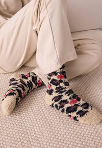 Chaussettes à motif léopard en beige, avec des accents multicolores en rouge et vert, fabriquées en tissu doux et texturé. Portées de manière décontractée avec un pantalon de couleur claire.