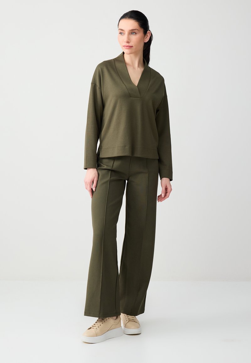 Vrouw staand, gekleed in een olijfgroene, lange mouw V-hals top, bijpassende wijde broek en beige sneakers, tegen een effen lichte achtergrond.