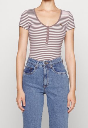 Persoon die een paars-wit gestreept henley-topje met korte mouwen draagt, ingestopt in een hoog getailleerde blauwe spijkerbroek met voorzakken en knoopsluiting.
