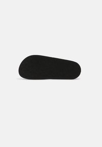 Semelle de chaussure noire avec un motif texturé de lettres G entrelacées sur un fond gris clair uni.