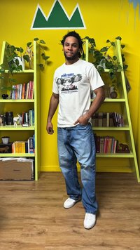 Maglietta bianca con stampa grafica, abbinata a jeans azzurri. Scarpe da ginnastica bianche. Parete gialla brillante con design di montagne verdi, piante su scaffali.