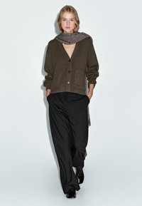 Bruine gebreide cardigan met twee voorzakken, knoopsluiting, gelaagd over een grijze sjaal, gecombineerd met losse zwarte broeken en zwarte schoenen.