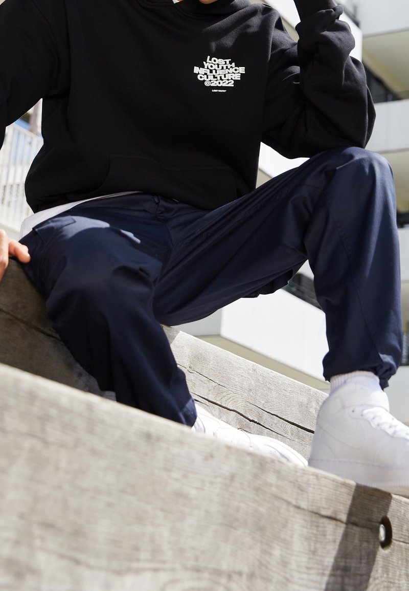 Personne portant un sweat à capuche noir avec un texte blanc, un pantalon bleu marine et des baskets blanches, assise sur des marches en bois en plein air.