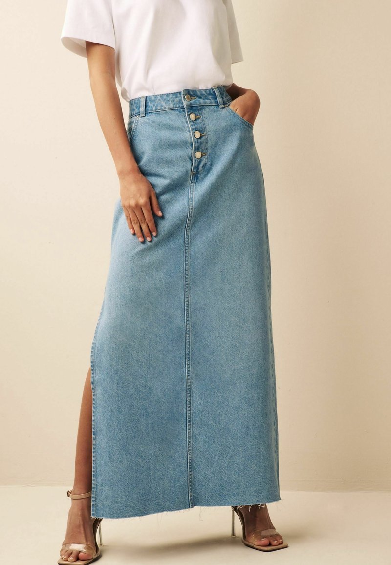 Femme portant une jupe longue en denim bleu clair taille haute avec des boutons sur le devant, une fente latérale, associée à un t-shirt blanc et des sandales à talons hauts transparentes.