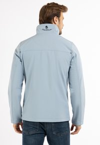 Svijetloplava softshell jakna s visokim ovratnikom, vidljivim šavovima, logotipom na leđima i dugim rukavima, u kombinaciji s tamnim trapericama.