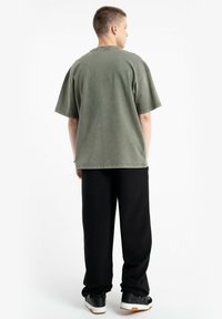Oliwkowa, oversize koszulka z krótkim rękawem, luźny krój oraz widoczne detale ściegów, w połączeniu z czarnymi szerokimi spodniami i trampkami.