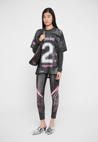 Svart oversized jersey med hvit nummer 2, kombinert med mønstrede, tettsittende leggings med rosa detaljer. Tilbehør inkluderer en blank, mørk håndveske og metalliske mules.