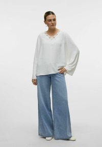 Hvid, langærmet top i struktureret stof med blonde detaljer ved halsudskæringen, parret med bredbenede blå denimbukser.