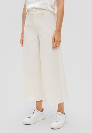 Persoon draagt offwhite wijde cropped broek met rauwe zomen en witte sneakers, staand tegen een effen grijze achtergrond.