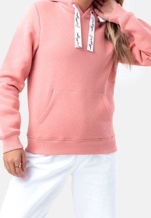 Žena nosící růžovou mikinu s kapucí s bílými šňůrkami a přední kapsou, v kombinaci s bílými kalhotami, ruka se dotýká vlasů.