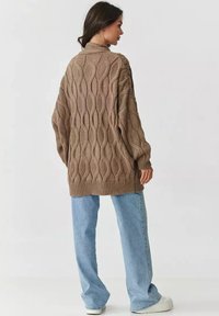Długi, oversize'owy brązowy sweter wykonany na drutach z prążkowanym dołem i wzorem w warkocze, w zestawieniu z jasnoniebieskimi spodniami o szerokich nogawkach i białymi trampkami.