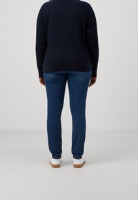 Navy tekstuuriga sviiter, millel on soonikkoes varrukad, koos tumeda sinise kitsaste teksade ja heledate pruunide talladega valgete tossudega, vaadatuna tagant.