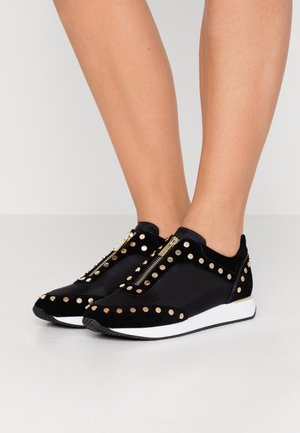 Sorte sneakers i en blanding af ruskind og stof, med gyldne nitter, lynlås-lukning og en hvid sål. Nærbillede af skoene.