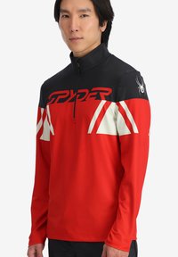 Pullover rosso con zip, spalle nere, dettagli bianchi e un logo. Leggero, texture liscia, maniche allungate e collo alto.