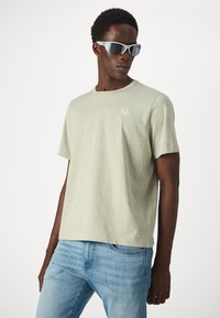 Hombre vistiendo una camiseta de manga corta verde claro con un pequeño emblema de flor y jeans azul claro, posando con gafas de sol deportivas blancas.