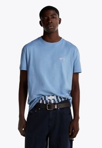 Maglietta di cotone azzurra chiara con un piccolo logo bianco sul lato sinistro del petto. Abbinata a pantaloni corti scuri e una cintura nera.