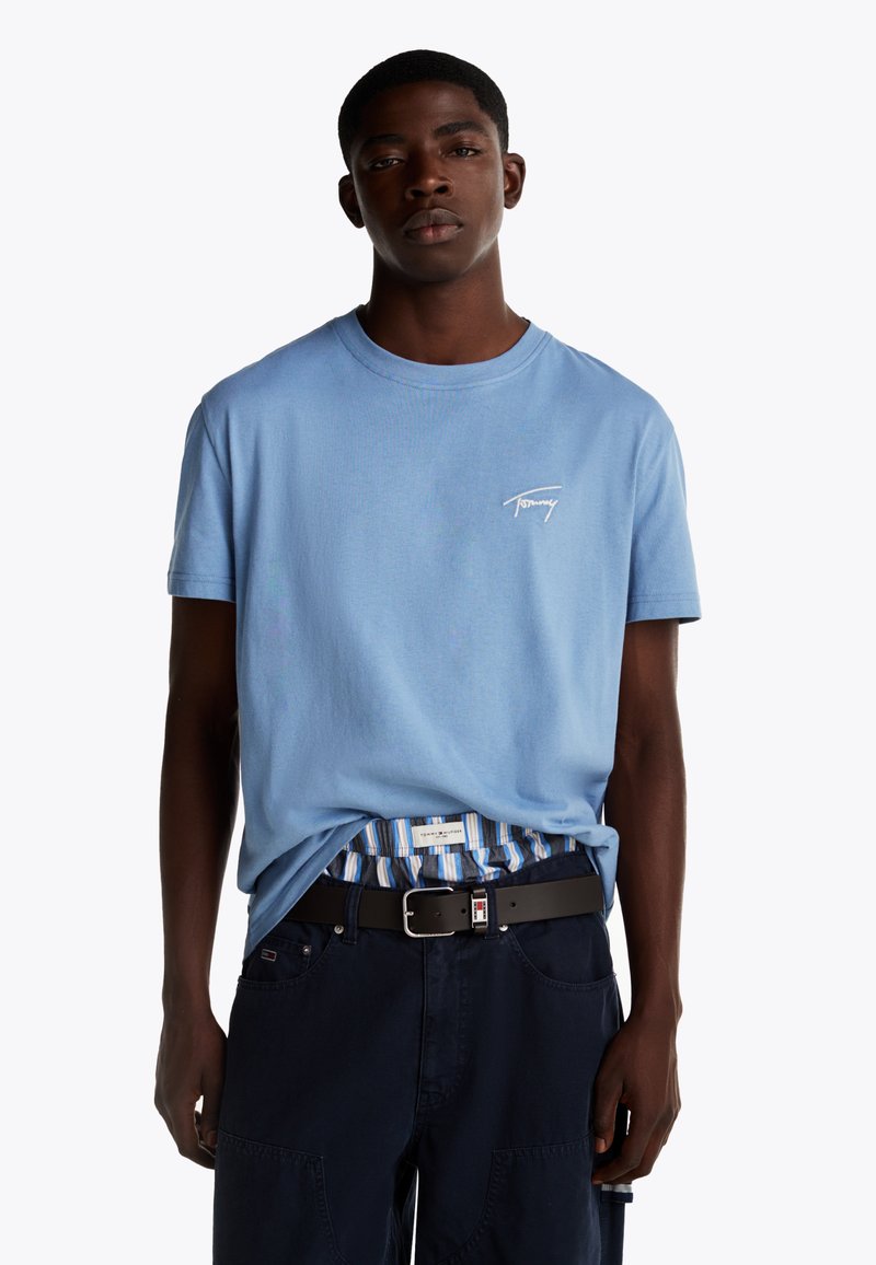 Maglietta di cotone azzurra chiara con un piccolo logo bianco sul lato sinistro del petto. Abbinata a pantaloni corti scuri e una cintura nera.