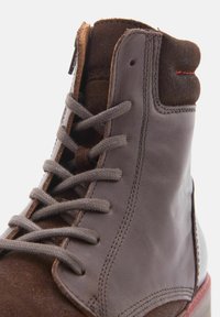 Bota de cuero marrón con cordones grises, con acentos de gamuza, punta redondeada y costuras rojas contrastantes a lo largo de la suela para darle detalle.