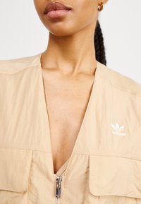 Beige lättviktsjacka med djup V-ringning, dragkedja och två fickor, med en vit Adidas-logotyp på vänster sida.