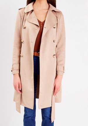 Femme portant un trench-coat beige à double boutonnage sur un haut marron et un jean bleu, debout devant un fond uni.