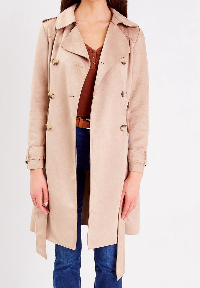 Femme portant un trench-coat beige à double boutonnage sur un haut marron et un jean bleu, debout devant un fond uni.