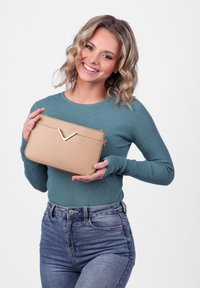 Hnědá kabelka z faux kůže s V-tvarovaným zlatým akcentem na přední straně, vybavená zipovým uzavřením a zaoblenými okraji. Drží ji osoba v tyrkysovém topu.