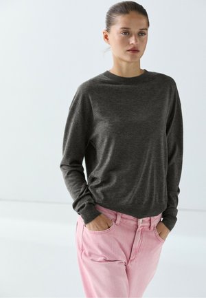 Junge Frau mit zurückgebundenem Haar trägt ein dunkelgraues Langarmshirt und hellrosa Jeans mit den Händen in den Taschen vor hellem, einfarbigem Hintergrund.