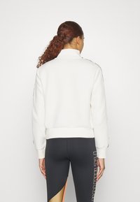 Sudadera blanca con cremallera y cuello alto, con puños acanalados, combinada con leggings negros que presentan un patrón geométrico multicolor.