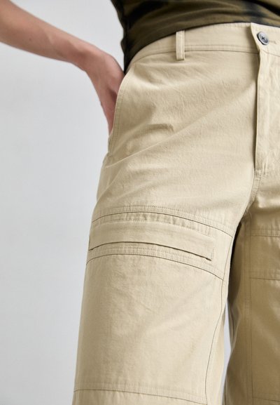 Beige cargo pants with a textured fabric, featuring side pockets and a waistband button closure. Designed with practical utility in mind.