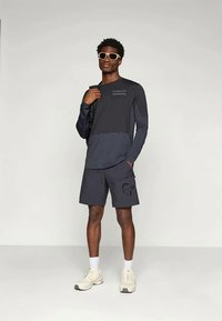 Tmavomodrá košeľa s dlhým rukávom a logom, skombinovaná s nohavicami zodpovedajúcej farby s grafickým potlačou. Model má na sebe slnečné okuliare a svetlú obuv.