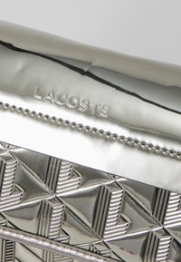 Srebrna metalna torbica z embosiranim geometričnim vzorcem, ki se ponaša z izstopajočim logotipom "LACOSTE" in elegantnim dizajnom. Teksturirani detajli na telesu.