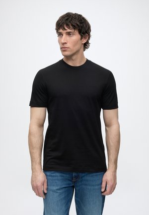 Homme aux cheveux foncés portant un t-shirt noir uni à manches courtes et un jean bleu, regardant vers la gauche sur un fond blanc.