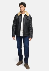 Schwarze Kunstlederjacke mit einem hellbraunen Schaffellkragen, ausgestattet mit Druckknöpfen und zwei Brusttaschen, kombiniert mit blauen Jeans und braunen Stiefeln.