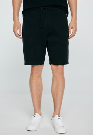 Mężczyzna w czarnych szortach z sznurkiem i białych trampkach, stojący na jednolitym jasnym tle.