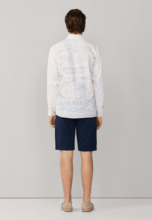 Hombre con camisa blanca de manga larga con texto y dibujos temáticos de playa en azul en la espalda, pantalones cortos azul marino y zapatos beige sin cordones.