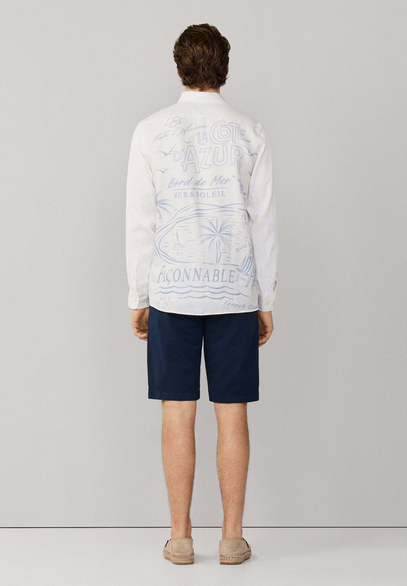 Hombre con camisa blanca de manga larga con texto y dibujos temáticos de playa en azul en la espalda, pantalones cortos azul marino y zapatos beige sin cordones.