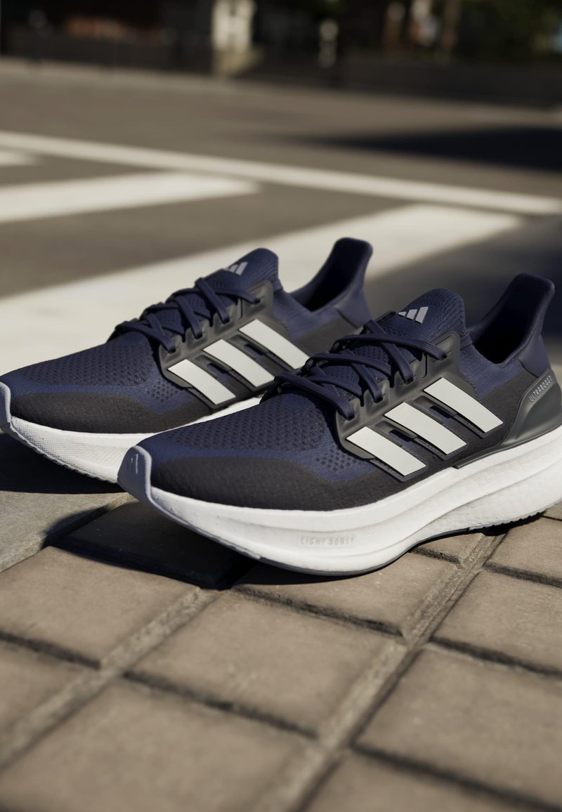 Zapatillas deportivas azul marino con parte superior de malla, diseño de tres rayas blancas, superficie texturizada, suela acolchada y detalles en gris en el talón.